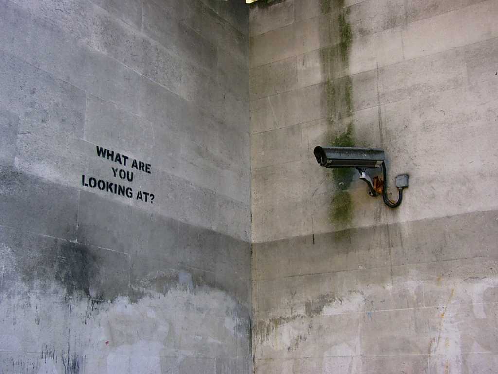 Foto av overvåkningskamera som ser på vegg med teksten ‘What are you looking at?’